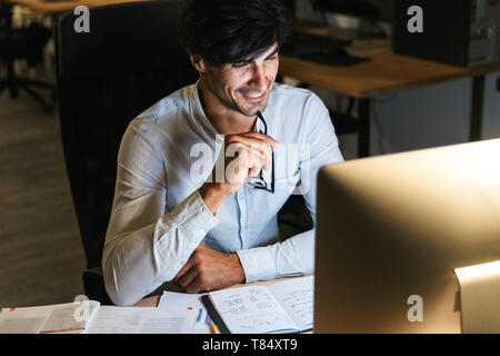 Smiling young man working at the office late at night, using computer Stock Photo
