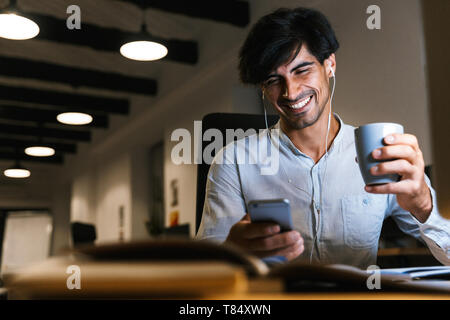 Smiling young man working at the office late at night, using computer, using mobile phone, drinking coffee Stock Photo