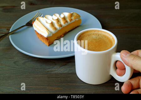 Hand Holding a Cup of Coffee with a Slice of Lemon Meringue Tart in Background Stock Photo