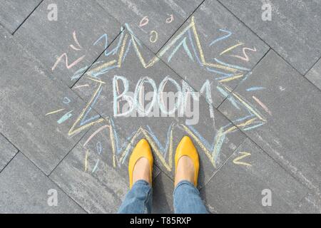 Boom written on gray sidewalk with women legs in yellow shoes, top view Stock Photo