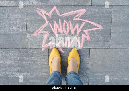 Wow written on gray sidewalk with woman legs in feet in yellow shoes, top view Stock Photo
