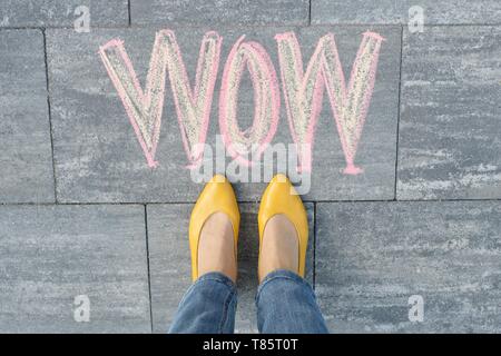 Wow written on gray sidewalk with woman legs in feet in yellow shoes, top view Stock Photo