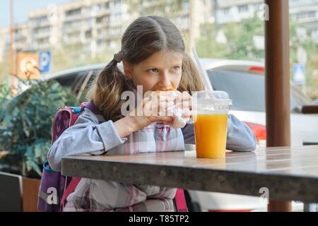 Portrait of girl student with backpack, eating burger with orange juice, background of outdoor fast food cafe. Stock Photo