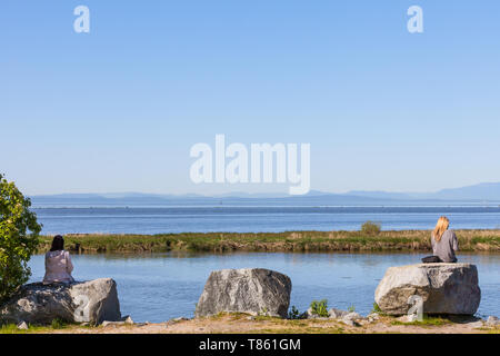 Two young women sitting on a rock looking across the sea to a distant Vancouver Island from Steveston British Columbia Stock Photo