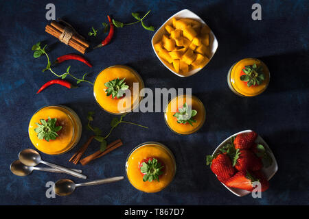 Traditional Italian sweet dessert Panna cotta with mango jelly served in jars and embellished with fresh real strawberrries and mint. Dark blue backgr Stock Photo