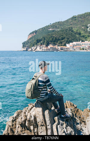 A tourist with a backpack alone sits on a cliff by the sea and looks into the distance at the view of Petrovac in Montenegro. Stock Photo