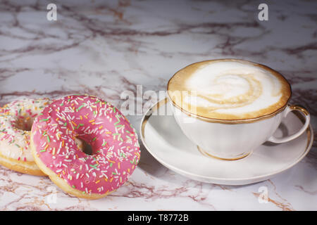 A cup of cappuccino coffee stands on a marble table next to two beautiful donuts Stock Photo