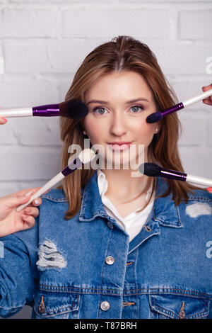 Studio photo of many hands with brushes doing make-up process to young caucasian beautiful woman on white background. Concept with many hands, lady is Stock Photo