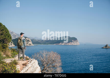 A tourist with a backpack on top of a cliff or hill next to the sea looks into the distance. Travel alone. Stock Photo
