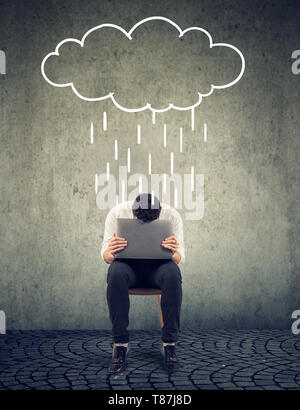Sad businessman sitting on a chair with laptop looking down with a rain cloud above Stock Photo