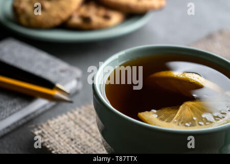 on a napkin from burlap there is a cup with black tea and two slices of lemon, on the background a plate of cookies, side of a gray notebook with pen  Stock Photo
