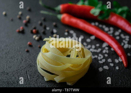 raw fettuccine paste on a dark background with fresh chili peppers, green branches of cilantro, coarse sea salt black and red peppercorns Stock Photo