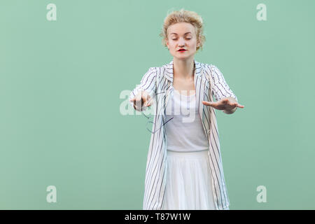 Portrait of blind young blonde woman in white shirt, skirt, and striped blouse standing, with closed eyes and trying to touch or find something. indoo Stock Photo