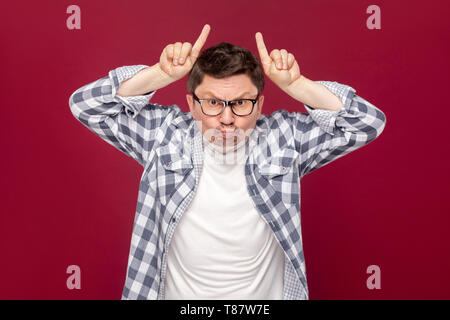 Portrait of angry middle aged business man in casual checkered shirt and eyeglasses standing with cow gesture horns on head and looking at camera. ind Stock Photo
