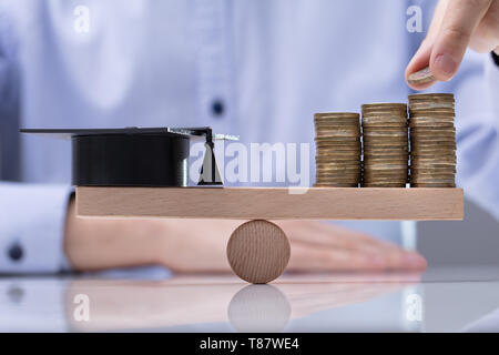 Man's Hand Placing Coin On Top Of The Stack Coins And Graduation Hat Over Seesaw Stock Photo