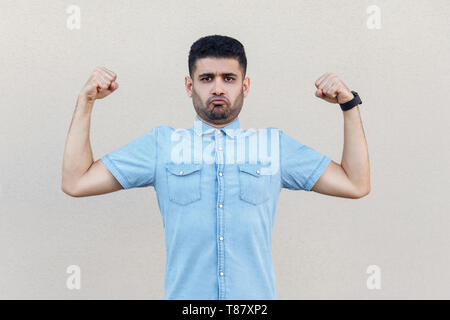 I am strong. Portrait of funny handsome young bearded man in blue shirt standing with raised arms and looking at camera with proud face. indoor studio Stock Photo
