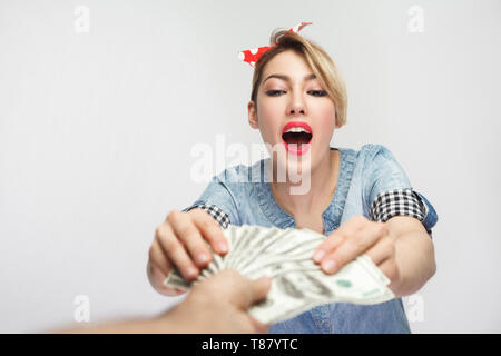 All for me! Portrait of wealthy amazed young girl in casual blue denim shirt with makeup, red headband standing and taking cash, mans hand giving mone Stock Photo