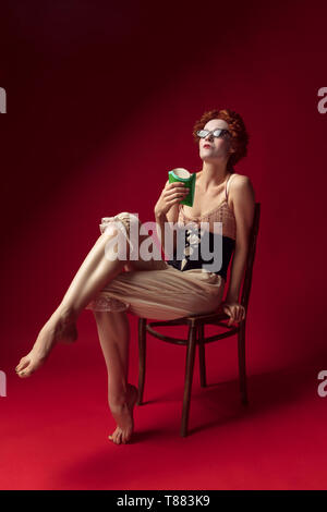 Medieval redhead young woman as a duchess in black corset, sunglasses and night clothes sitting on red background eating fried potato. Concept of comparison of eras, modernity and renaissance. Stock Photo