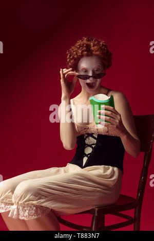 Medieval redhead young woman as a duchess in black corset, sunglasses and night clothes sitting on red background eating fried potato. Concept of comparison of eras, modernity and renaissance. Stock Photo