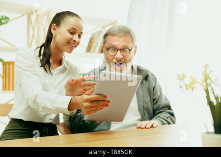 A daughter or granddaughter spends time with the grandfather or senior man. Family or fathers' day, emotions and happieness. Lifestyle portrait at home. Girl taking care about dad. Using a tablet. Stock Photo