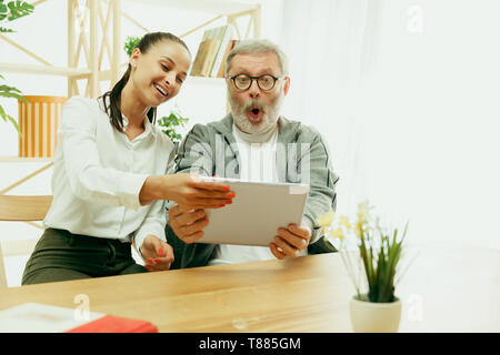 A daughter or granddaughter spends time with the grandfather or senior man. Family or fathers' day, emotions and happieness. Lifestyle portrait at home. Girl taking care about dad. Using a tablet. Stock Photo