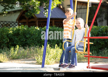 Cute boy pushing little girl on swings outdoors Stock Photo