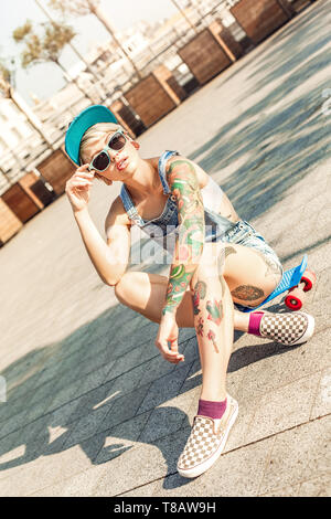 Skateboarding. Alternative girl skater in cap and sunglasses sitting on penny board on the city street looking aside cool Stock Photo