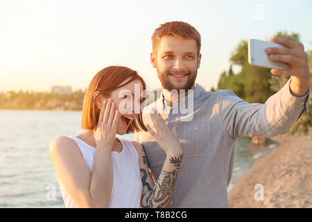 Happy couple taking selfie with engagement ring near river on sunny day Stock Photo