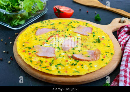 Omelet with ham, cheese, greens on a wooden board on a black background.  Fritata - a delicious, healthy breakfast. Stock Photo