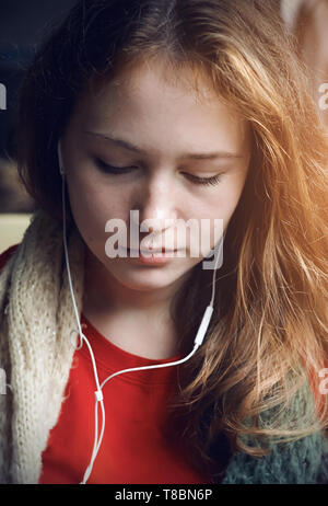 A long-haired beautiful girl in a red t-shirt rides in public transport near the window, from which the sunlight shines, and listens to music in headp Stock Photo