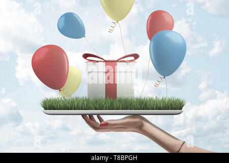 Side view of woman's hand holding digital tablet with green grass on screen, gift box standing on top and colorful balloons flying around. Stock Photo
