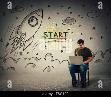 Creative young man freelancer working on laptop seated on a chair. Cheerful positive guy playing video games on his notebook computer as rocket ship s Stock Photo