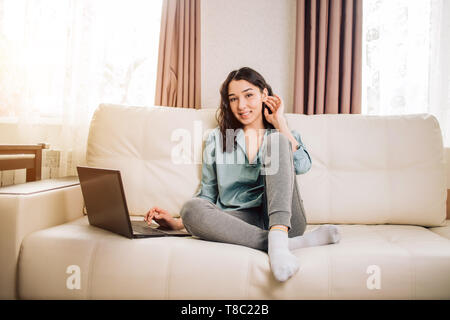 Relaxed female freelancer touching her dark wavy hair during working with laptop. Indoor portrait of cute young lady in shirt and pants sitting on whi Stock Photo