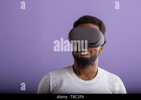 Young dark skinned man dressed in casual outfit wearing mobile virtual reality headset for playing 3D game isolated on purple background. VR technolog Stock Photo