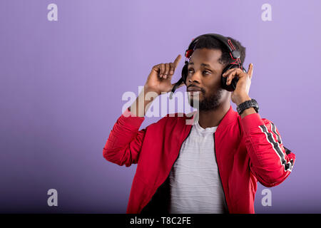 Studio portrait of handsome young black man in headphones, enjoying listening to music and dancing on lilac background. Professional african american  Stock Photo