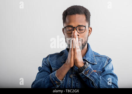 Closeup of young black man praying with hands clasped together, asking the God for good luck, success, health, looking up, isolated gray studio wall.  Stock Photo