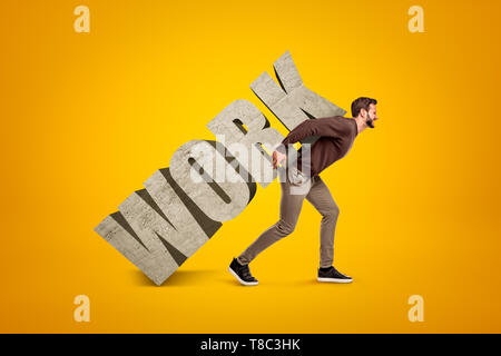 Young man in casual clothes carrying heavy WORK sign on his back on yellow background Stock Photo