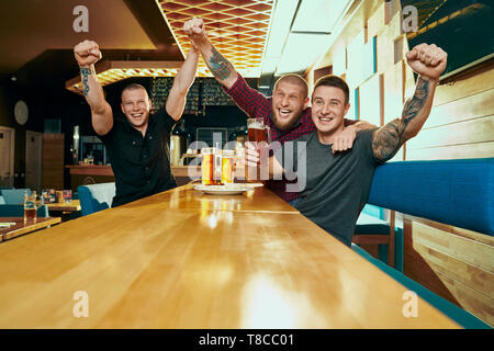 Front view of three male fans watching football, cheering and resting on weekends in pub. Cheerful men sitting at table, drinking beer and laughing in bar. Concept of happiness and leisure. Stock Photo