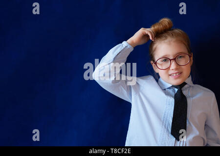 Little confused schoolgirl scratching head. Bewildered preschool girl in school uniform and tie. Schoolchild in glasses on dark blue background. Elementary grade student, kindergarten pupil Stock Photo