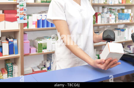 Unrecognizable worker of drugstore standing at counter at workplace. Woman scanning barcode of medicament with special scanner. Pharmacist in white unifrom holding white medical box. Stock Photo