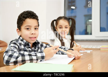 Pupil of primary school sitting at desk, holding pencils, looking at camera, posing. Cheerful boy and girl, schoolchildren learning, writing in copybook in classroom. Stock Photo