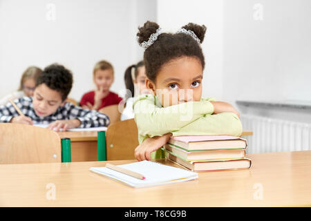Pretty schoolgirl sitting at desk with copybook and pen in classroom, leaning on books. Cute child posing, looking at camera. Group of pupils sitting at tables in class, writing.  Stock Photo