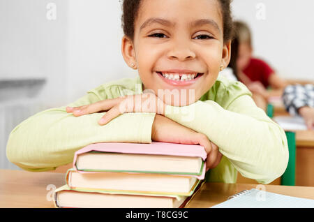Cute, pretty schoolgirl sitting at desk of school, leaning on books, smiling, lookling at camera. Portrait of happy, cheerful pupil in classroom of primary school. Stock Photo