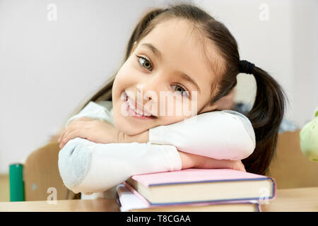 Portrait of pretty, funny schoolgirl with pigtails in classroom. Pupil sitting at desk, leaning with hands on books, smiling. Child looking at camera, posing. Stock Photo
