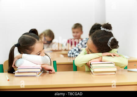 Young, pretty schoolgirls, sitting in classroom at desk and leaning on stack of books. Classmate sleeping on books. Schoolchildren, pupils studying in classroom. Stock Photo
