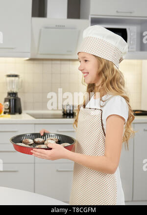 Charming, cute little girl holding metal square baking form with homemade chocolate cookies. Cheerful, happy teenager wearing in chef hat and apron, smiling, looking away. Stock Photo