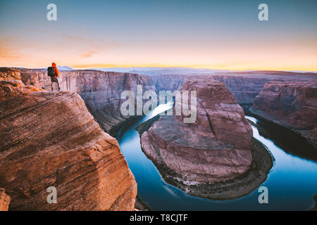 A male hiker is standing on steep cliffs enjoying the beautiful view of Colorado river flowing at famous Horseshoe Bend at sunset, Arizona, USA Stock Photo