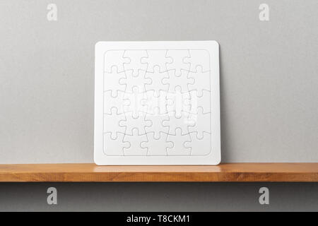 Design concept - front view of white square jigsaw puzzle on bookshelf and grey wall for mockup Stock Photo