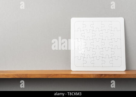 Design concept - front view of white square jigsaw puzzle on bookshelf and grey wall for mockup Stock Photo