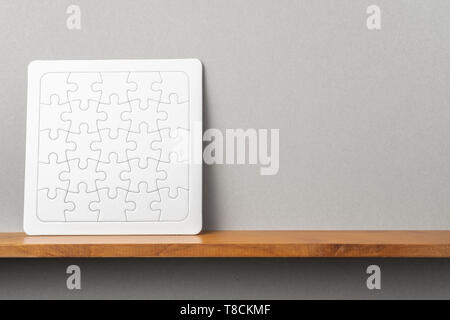 Design concept - front view of white square jigsaw puzzle on bookshelf and grey wall for mockup Stock Photo